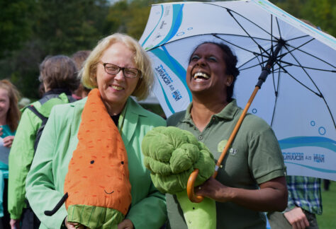 A woman in green holds a large stuffed carrot while another woman in green laughs holding a large stuffed broccoli.
